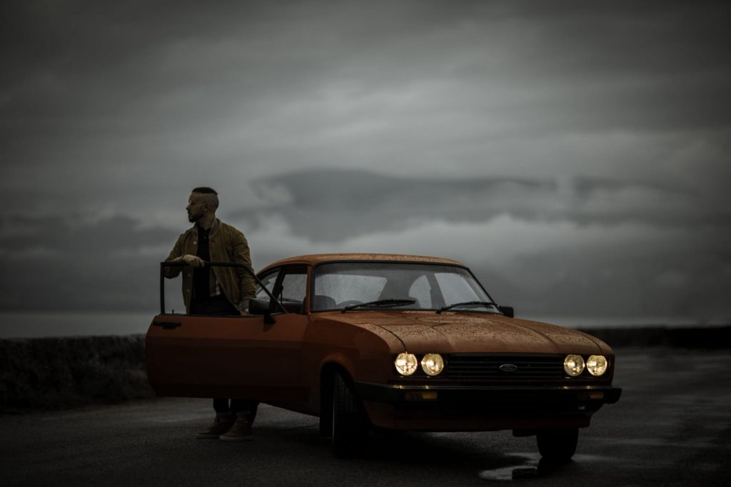 Gabriele (aka Gangalistics) stands behind the open door of an orange car. A grey sky looks over him.