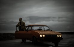 Gabriele (aka Gangalistics) stands behind the open door of an orange car. A grey sky looks over him.