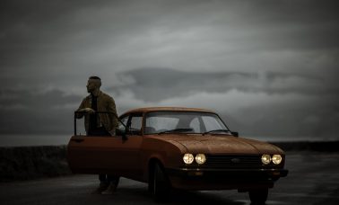 Gabriele (aka Gangalistics) stands behind the open door of an orange car. A grey sky looks over him.