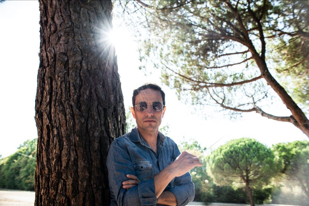 Italian electronica star AGA teams with Liverpool synthwave artist for new single AGA leans against a tree wearing a blue jacket. He wears sunglasses and the sun shines in the background.