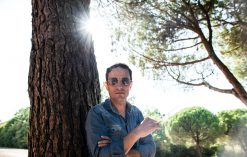 AGA leans against a tree wearing a blue jacket. He wears sunglasses and the sun shines in the background.