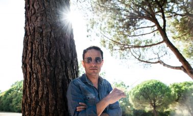 AGA leans against a tree wearing a blue jacket. He wears sunglasses and the sun shines in the background.