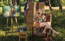 A performer sitting outside perched on a chair and eating a packet of crisps. Around her are other bits of furniture in a forest setting.