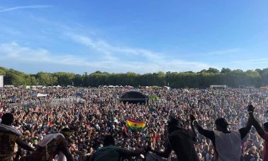 A band take a bow in front of a packed festival crowd underneath a blue sky.