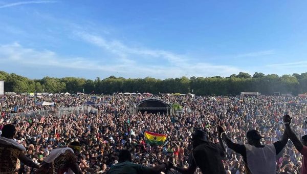 A band take a bow in front of a packed festival crowd underneath a blue sky.